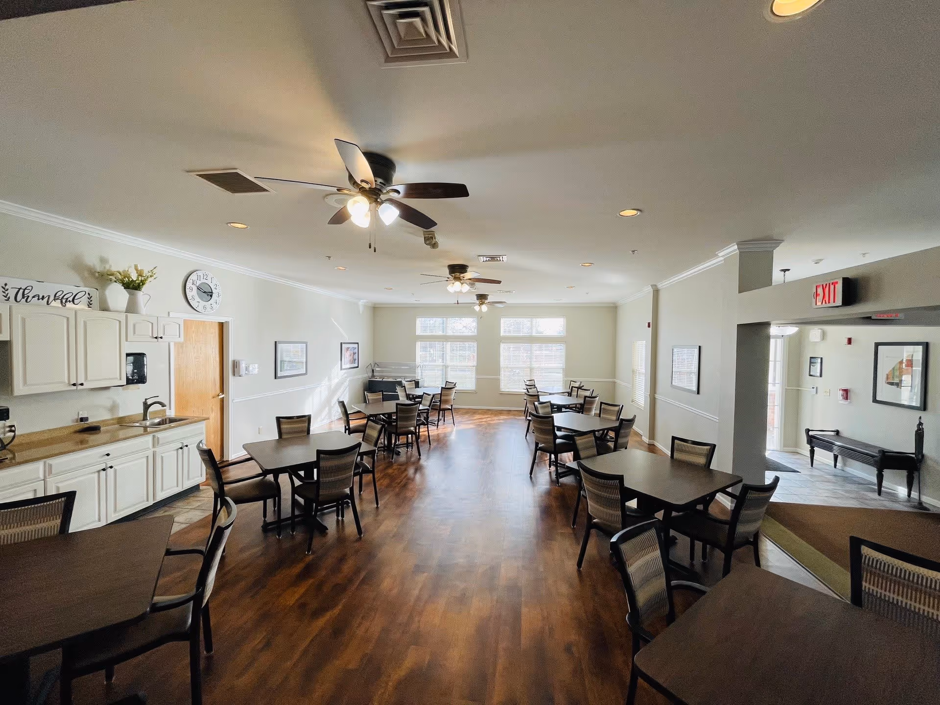 A bright dining room with multiple dark wood tables and chairs arranged neatly on a wooden floor. The room has white walls, ceiling fans with lights, large windows letting in natural light, and a small kitchenette area with cabinets and a sink on the left side. There is an exit sign above a doorway on the right side.