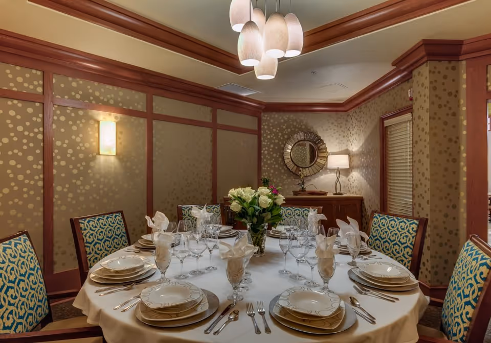 Round dining table set for a meal with plates, glassware, napkins and a floral centerpiece in a warmly lit private dining room.