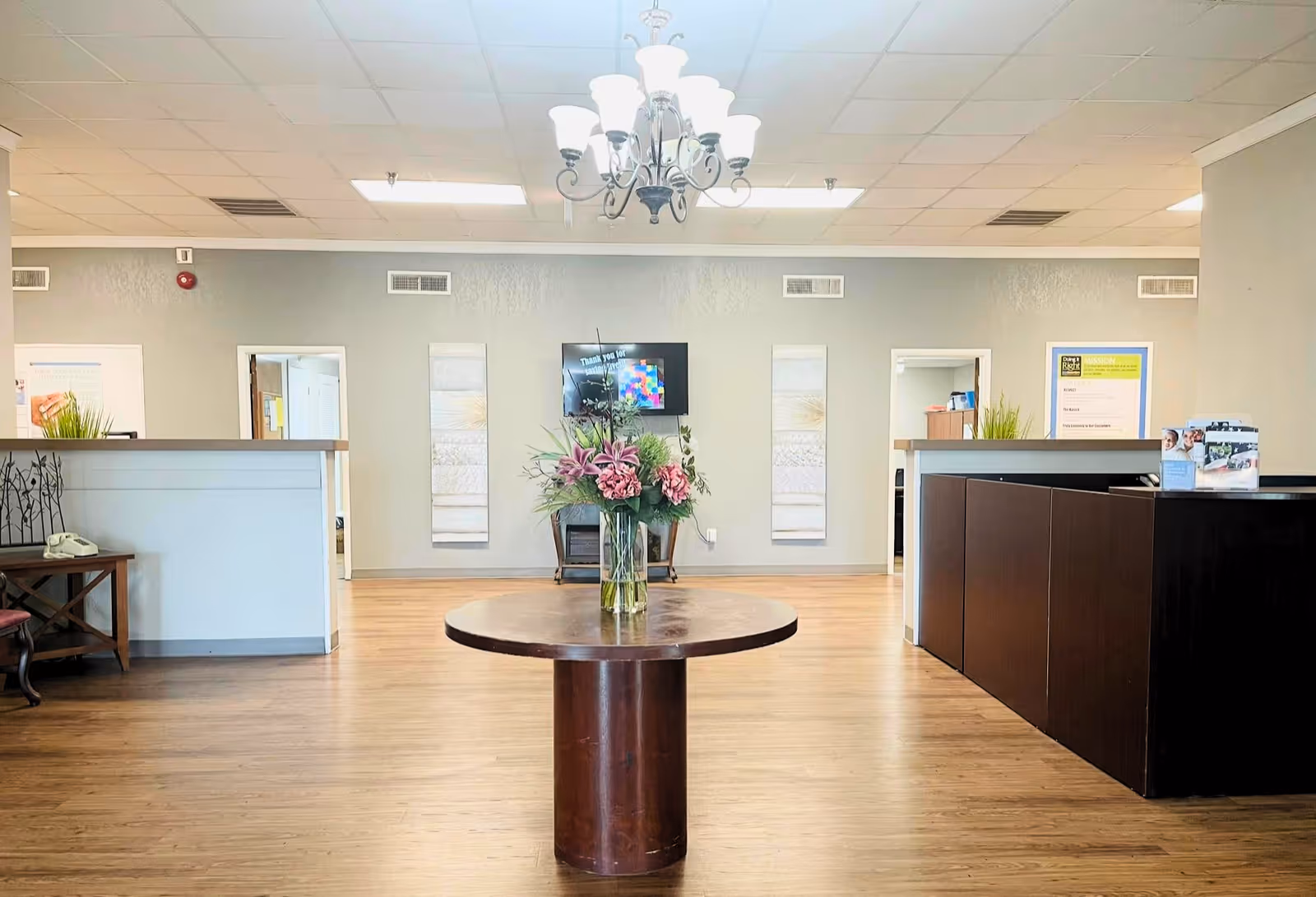 Interior view of a reception area in a senior living facility with a round wooden table in the center holding a vase of pink flowers. There are two reception desks on either side, a chandelier hanging from the ceiling, and a wall-mounted TV with decorative panels on the back wall.