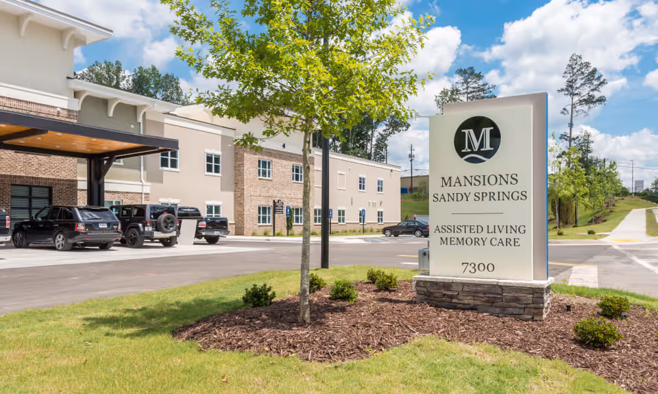 Exterior view of Mansions Sandy Springs assisted living and memory care facility with a large sign in the foreground displaying the facility name and address. Several cars are parked near the building under a covered entrance, with trees and a clear blue sky in the background.