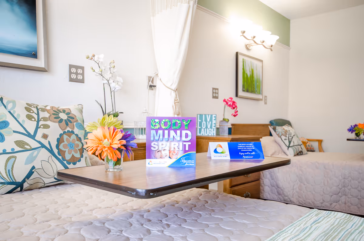 A bright and cozy room with two beds featuring floral and patterned pillows. A wooden overbed table is positioned between the beds, holding a small vase with colorful flowers and two decorative signs, one reading 'BODY MIND SPIRIT' and the other 'LIVE LOVE LAUGH'. The walls are light-colored with framed artwork and a wall-mounted light fixture above one bed.
