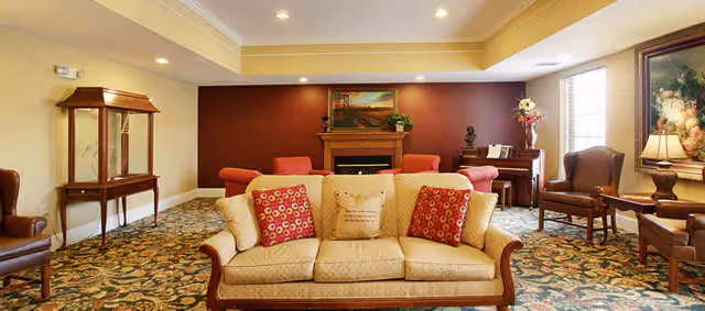 Cozy senior living common room with a sofa and chairs arranged around a fireplace, piano, and decorative artwork.