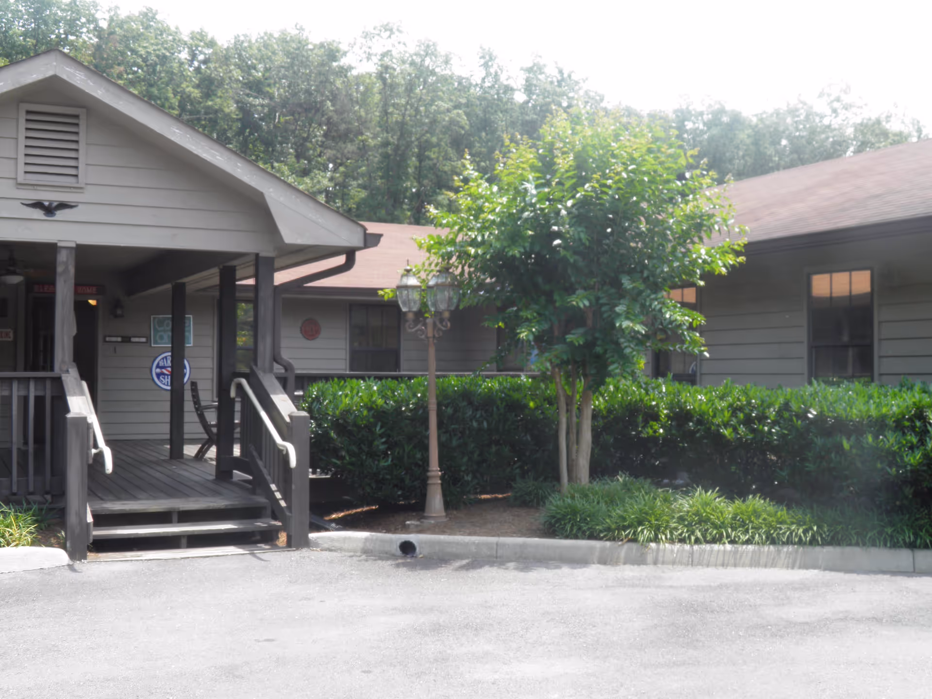 Exterior view of a single-story building with a covered porch entrance, surrounded by greenery including bushes and a small tree. There is a vintage-style street lamp near the entrance and a paved driveway in front.
