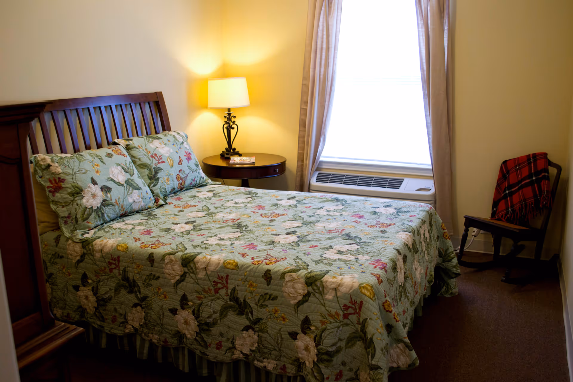 A cozy bedroom with a wooden bed frame and a floral patterned bedspread and pillows. There is a round wooden nightstand with a lamp on it next to the bed. A window with beige curtains and an air conditioning unit below it is on the wall. A wooden chair with a red plaid blanket draped over it is in the corner of the room.