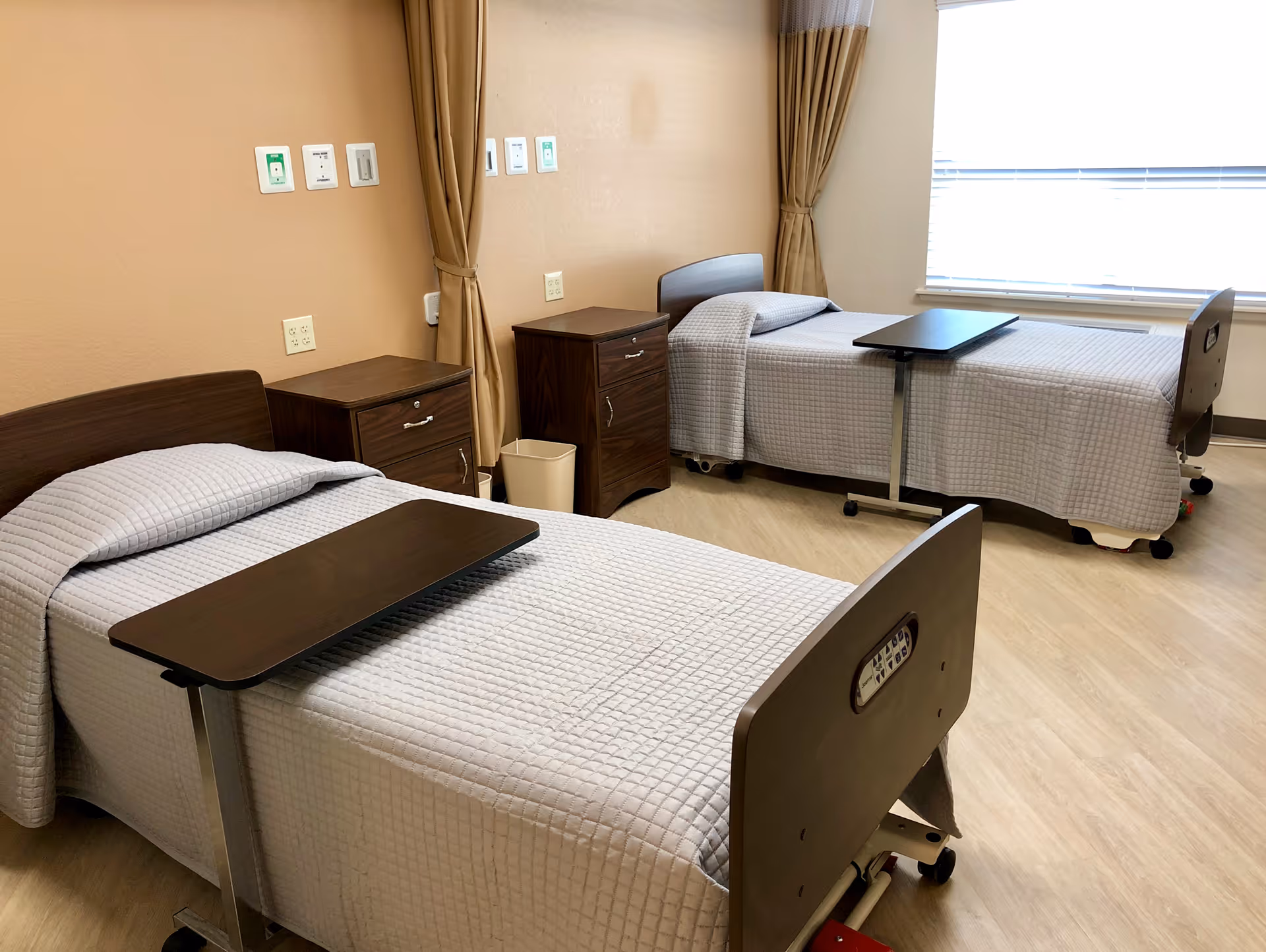 A clean and well-lit room with two hospital-style beds, each covered with a light gray quilted blanket and equipped with an overbed table. There are two wooden nightstands between the beds, a beige trash bin, beige curtains, and a window with blinds letting in natural light. The walls are painted a light tan color and the floor has a light wood finish.
