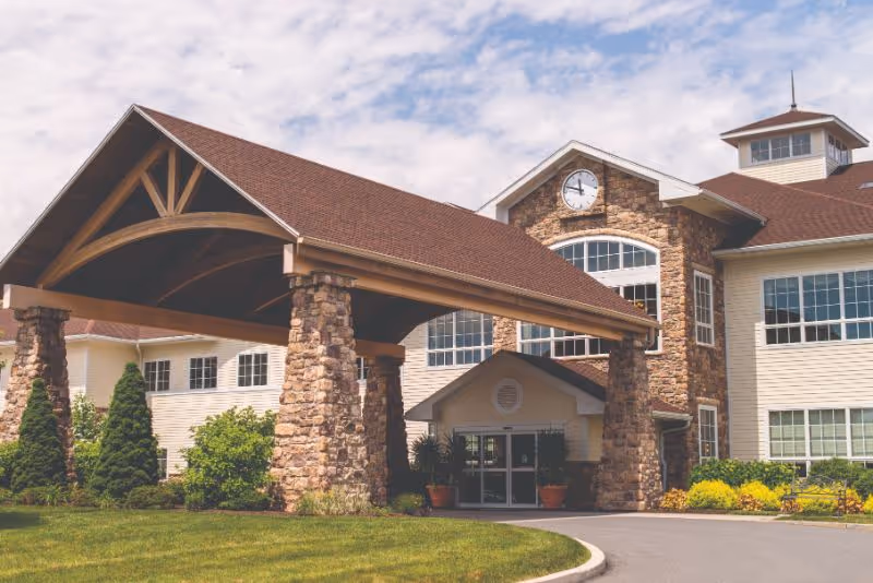 Exterior view of The Heritage Of Green Hills senior living facility showing a large covered entrance with stone pillars, a clock above the main entrance, multiple windows, and well-maintained landscaping with green shrubs and trees under a partly cloudy sky.