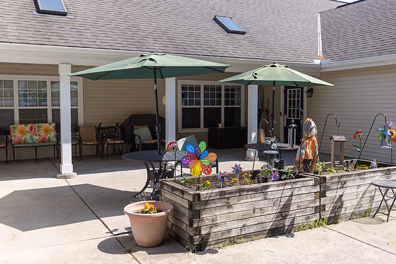 Sunlit outdoor patio courtyard with green umbrellas, tables and chairs, raised wooden planters and colorful pinwheel decorations in front of a residential building.