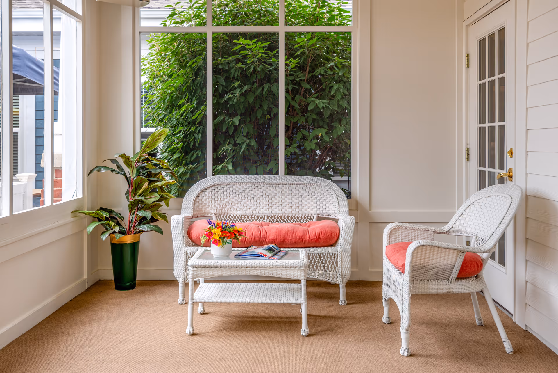 A bright enclosed porch with white wicker sofa and chair with red cushions, a small white wicker table holding a flower arrangement and magazine, and a potted plant by large windows.