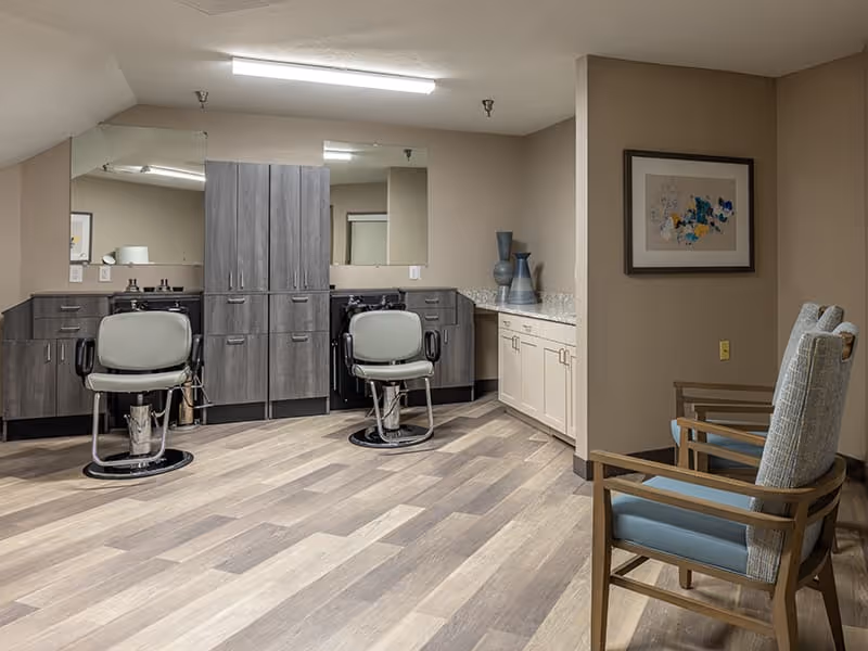 Interior view of a salon area in a senior living facility with two gray salon chairs in front of a large mirror and cabinetry. The room has wood-patterned flooring, beige walls, and a small seating area with two wooden chairs with blue cushions. Decorative vases and framed artwork are on the countertop and wall.