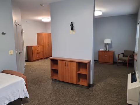 Interior view of a senior living facility room with gray walls and carpeted floor. The room features wooden furniture including a cabinet with shelves in the center, a dresser and wardrobe in the background, and a small table with a lamp and chair near a window. A hospital-style bed with white sheets is partially visible on the left side.