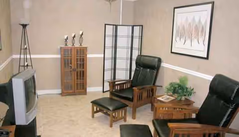 Cozy lounge with green recliners, wood tables, a folding screen, cabinet, plant, and TV.