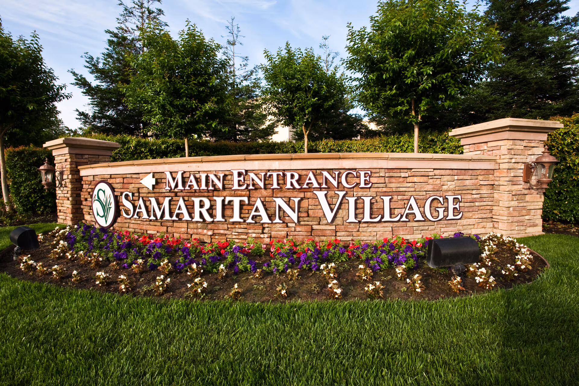 A large stone sign with the text 'Main Entrance Samaritan Village' surrounded by a flower bed with colorful flowers and green grass, with trees and bushes in the background.