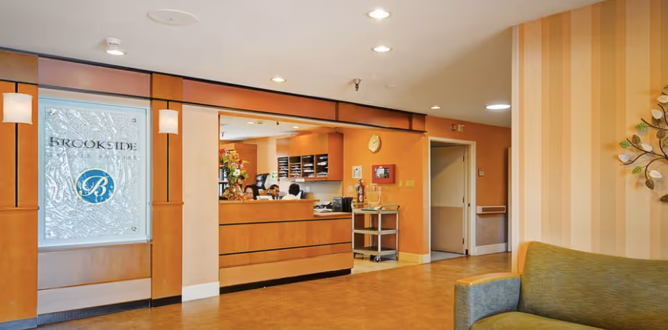 Reception area of Brookside Skilled Nursing Hospital with a wooden front desk, staff working behind the desk, warm orange walls, a green couch in the foreground, and decorative wall art on the right.