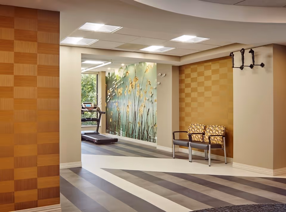 Interior view of a hallway in Oak Brook Healthcare Centre featuring patterned walls in shades of brown, two chairs with patterned cushions against the wall, a coat rack with three hooks, and a treadmill near a large window with a nature-themed mural of tall grasses on the wall.