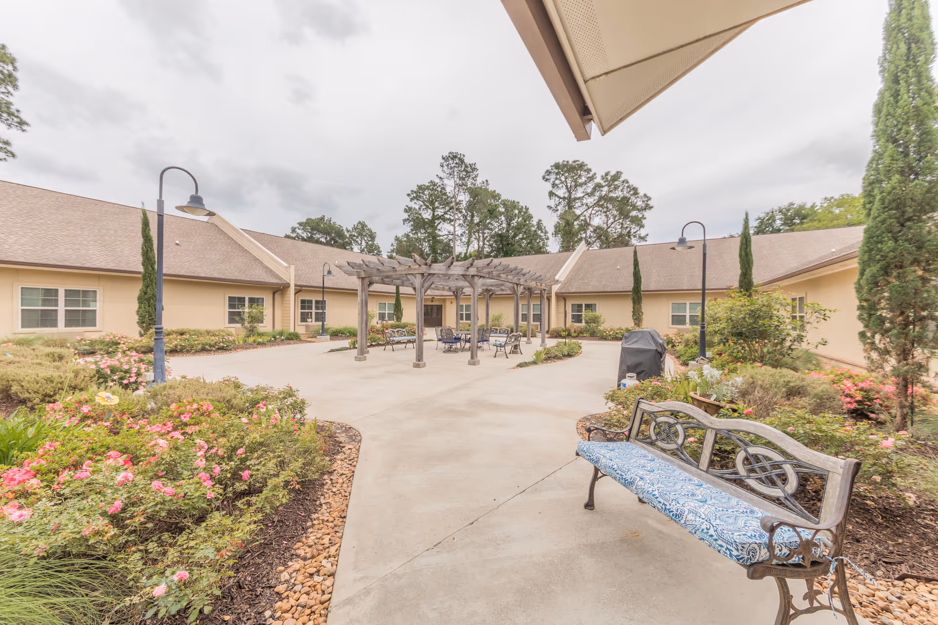 Outdoor courtyard area at Cedar Crest Memory Care featuring a paved walkway, a wooden pergola with seating underneath, a cushioned bench, garden beds with blooming flowers and shrubs, tall trees, and beige single-story buildings surrounding the courtyard under a cloudy sky.