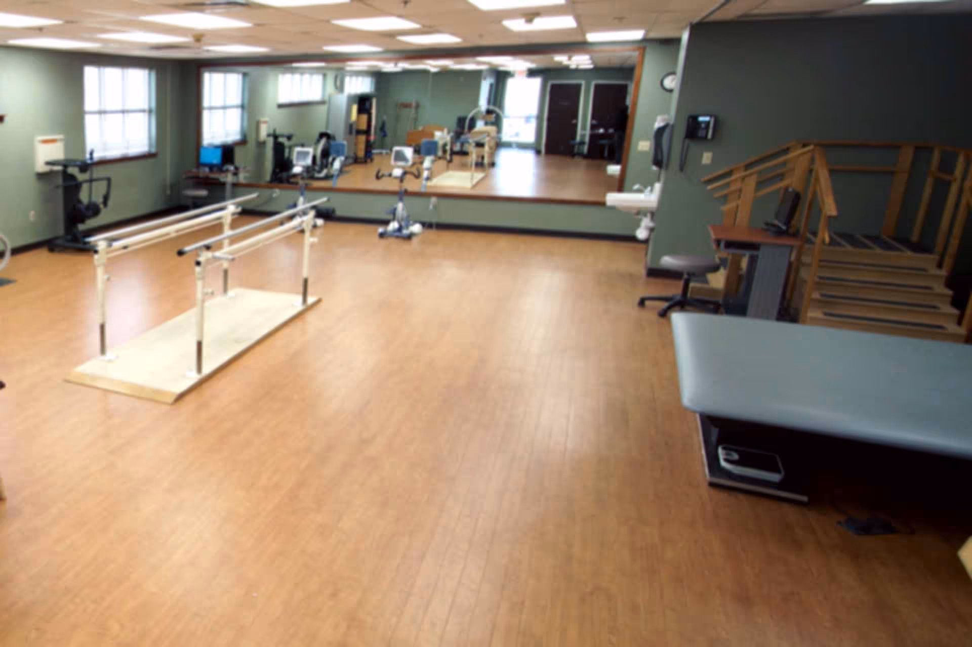 A spacious rehabilitation room with wooden flooring, parallel bars for walking exercises, exercise equipment, a treatment table, a chair, a small staircase with handrails, and a large mirror on the far wall reflecting the room.