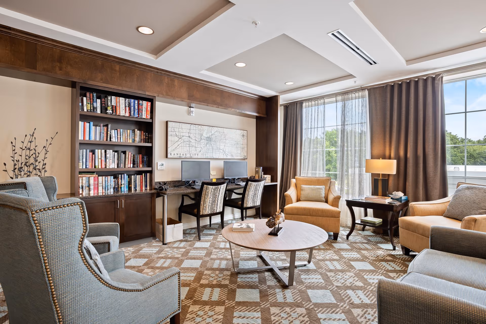 A cozy living room area with two blue armchairs and two beige armchairs arranged around a round wooden coffee table. Behind the seating area is a built-in bookshelf filled with books and a desk with two computer monitors and chairs. Large windows with sheer and dark curtains allow natural light to fill the room.