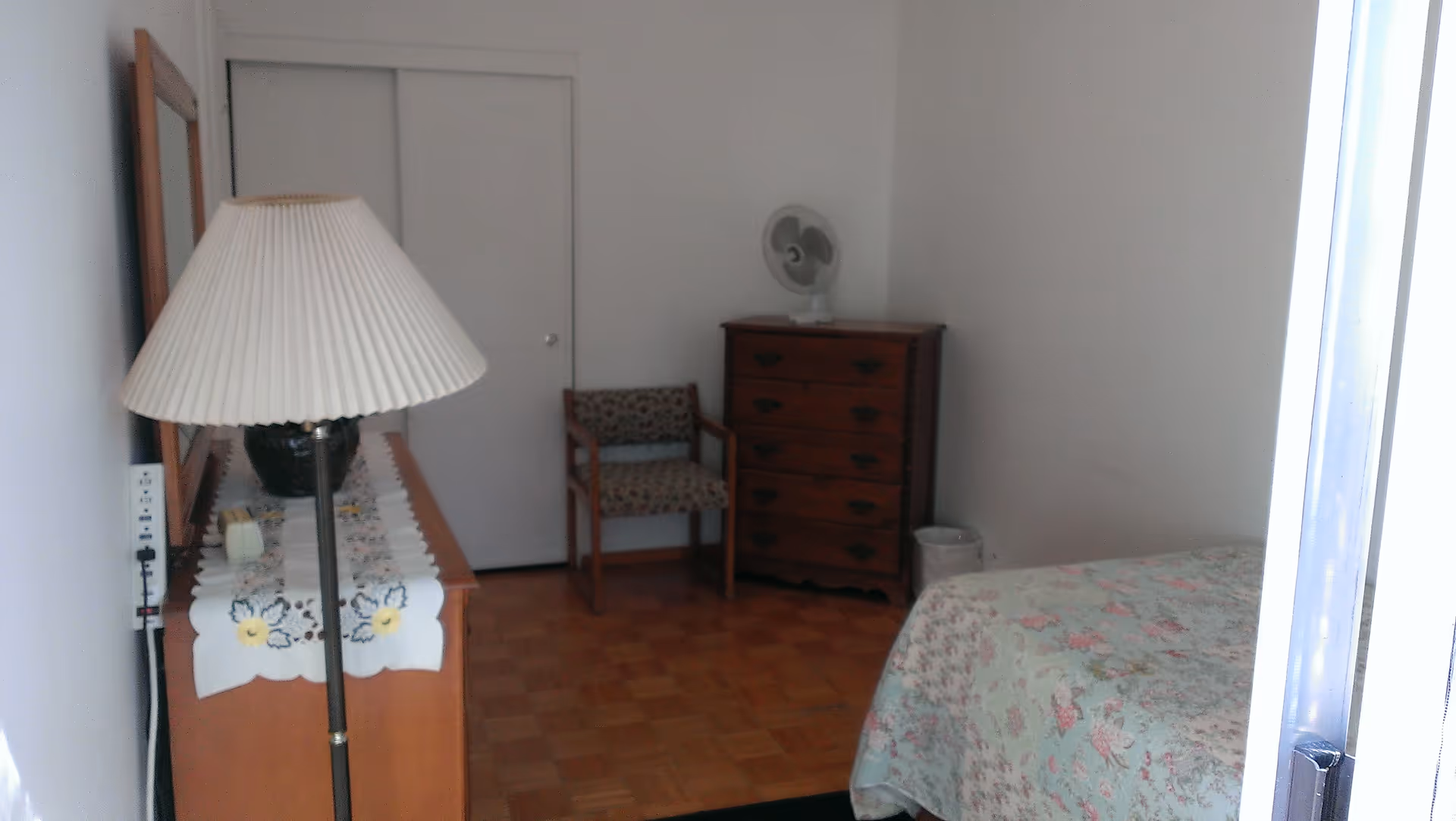 Small bedroom with a bed covered by a floral quilt, a wooden dresser, chair, floor lamp, and a sliding closet door.