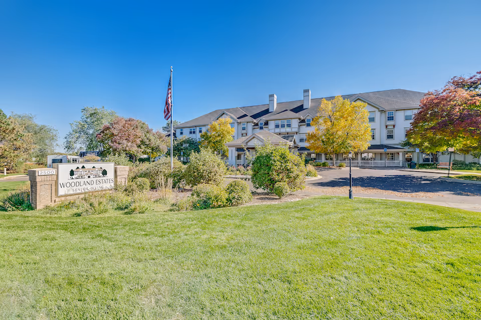 Exterior view of Woodland Estates Senior Living facility with a large green lawn, trees with autumn foliage, an American flag on a flagpole, and a sign displaying the facility's name and address 2500.