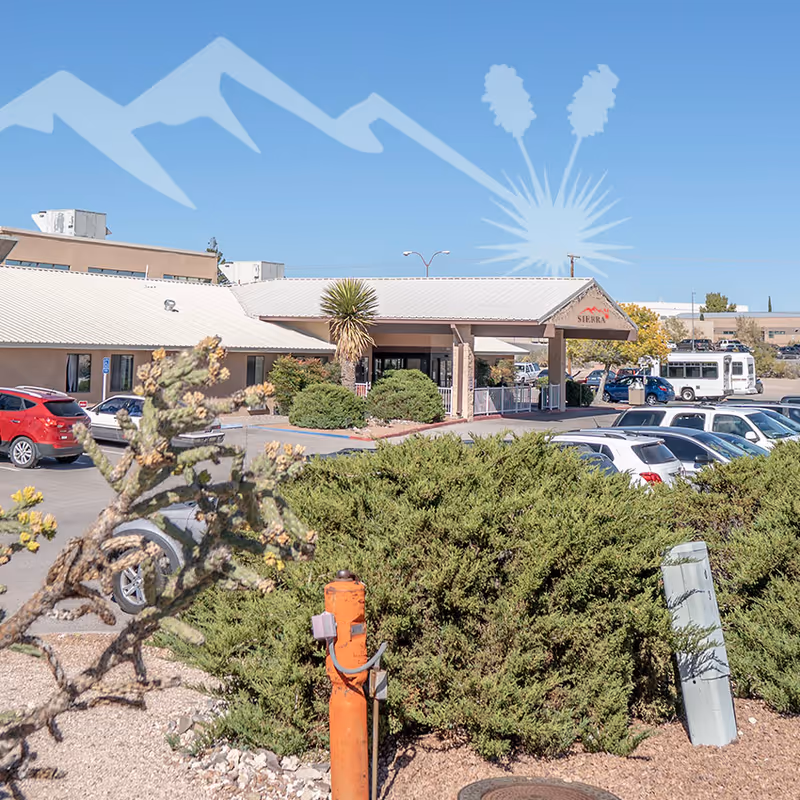 Exterior view of Sierra Hill Assisted Living facility showing the entrance with a covered drop-off area, surrounded by bushes and a parking lot with several cars under a clear blue sky.