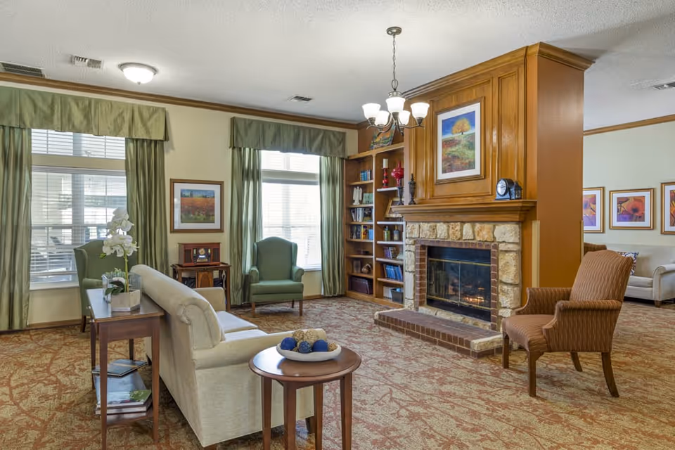 A cozy living room area with a stone fireplace, wooden mantel, and built-in bookshelves. The room features a beige sofa, two green armchairs near windows with green curtains, and a brown armchair. A round wooden side table with decorative balls and a rectangular table with a white orchid plant are also present. The walls are light-colored with framed artwork, and a chandelier hangs from the ceiling.