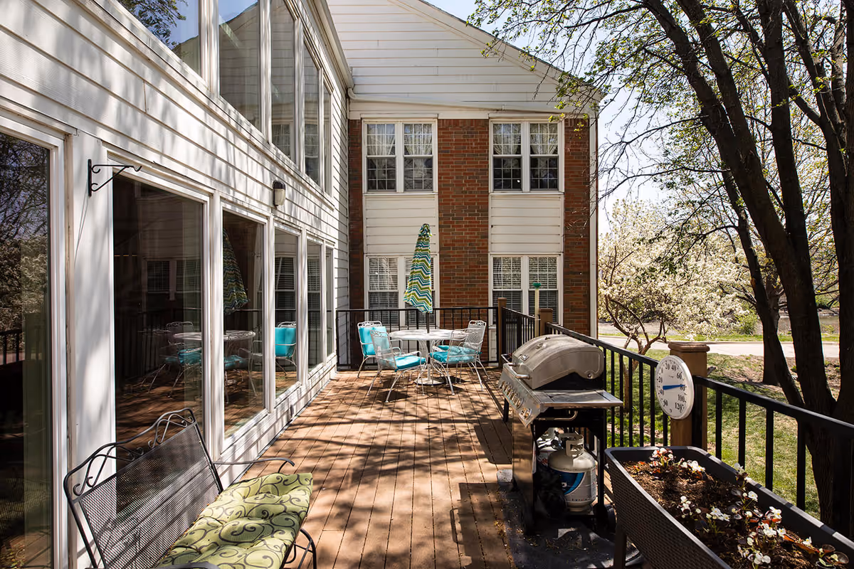 Outdoor patio area at Lexington Park Assisted Living of Topeka featuring a wooden deck with a metal bench with green cushion, a round table with four turquoise cushioned chairs and a striped umbrella, a large gas grill, a planter box with flowers, and a thermometer attached to the railing. The patio is adjacent to a building with large windows and surrounded by trees and greenery.