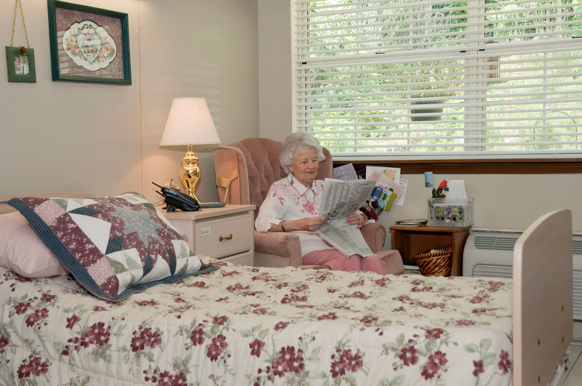 An elderly woman sitting in a pink armchair reading a newspaper in a cozy bedroom with floral bedding and a quilted pillow on the bed. The room has a nightstand with a lamp and a phone, a window with blinds, and some decorations on the walls.