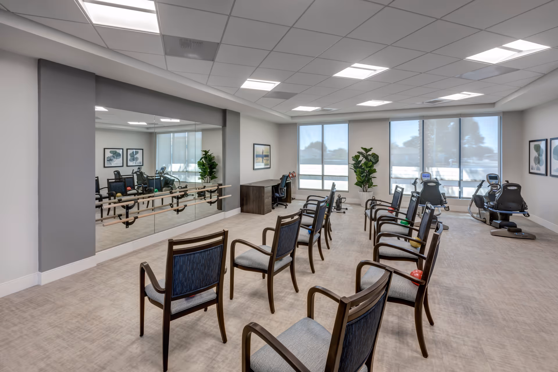 Bright exercise and therapy room with rows of chairs, wall mirrors, and stationary fitness equipment.