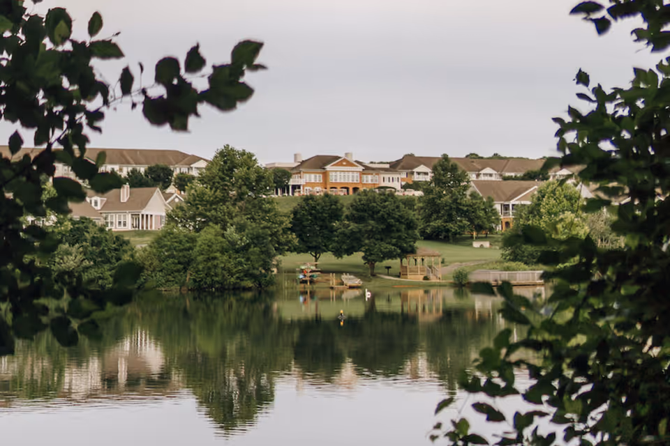 View of a senior living facility named The Summit with multiple buildings surrounded by green trees and a calm lake in the foreground reflecting the buildings and trees.