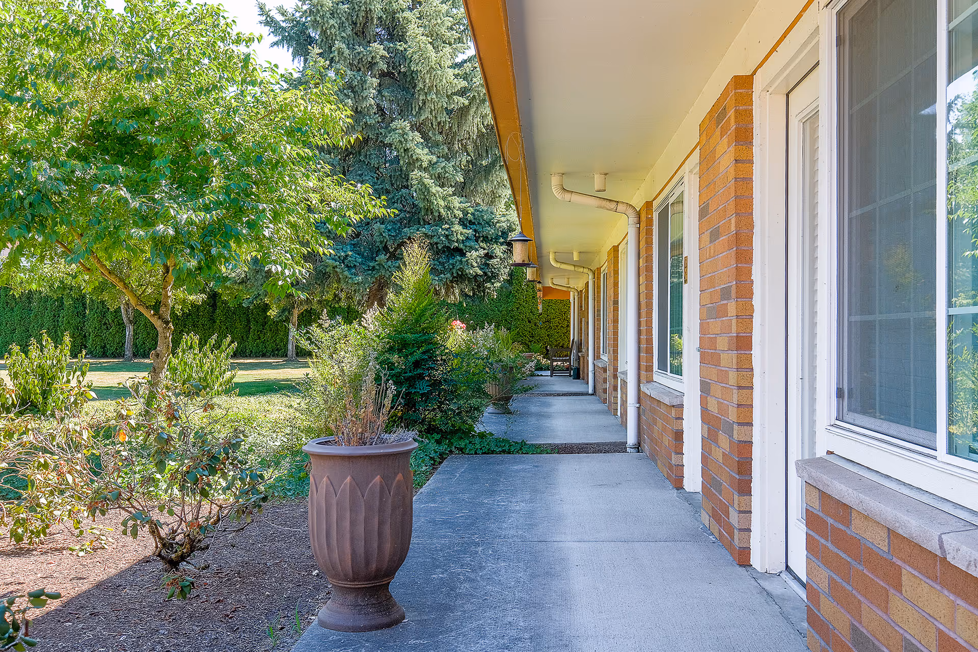 A covered outdoor walkway alongside a brick building with windows and doors. The walkway is bordered by a garden area with various green plants, bushes, and a tree. There is a large decorative planter on the edge of the walkway. The scene is bright and sunny with clear skies.
