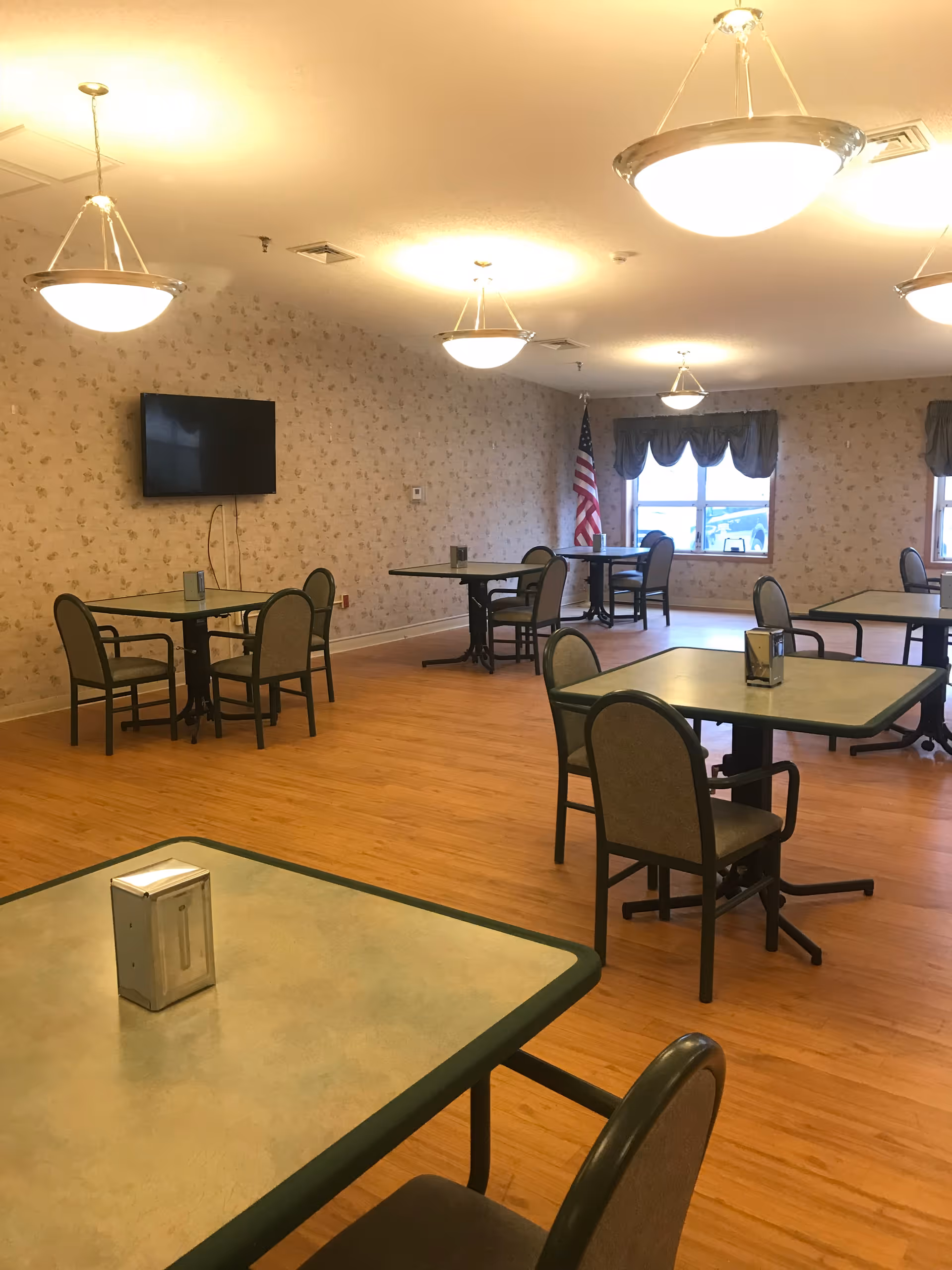 A dining room with several square tables and chairs arranged neatly on a wooden floor. The walls have a light floral wallpaper, and there are multiple ceiling lights providing warm illumination. A flat-screen TV is mounted on one wall, and an American flag is positioned near a window with blue curtains.