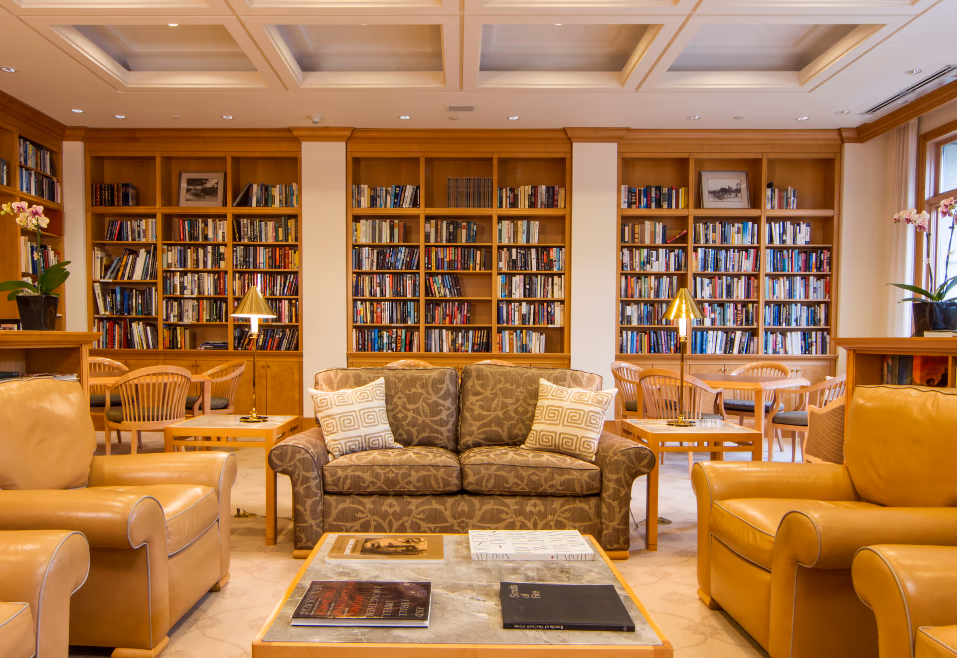 A cozy library room with wooden bookshelves filled with books along the back wall. In front of the shelves, there is a patterned fabric sofa with two decorative pillows, flanked by two tan leather armchairs. Two small tables with brass lamps are positioned beside the sofa. There are also wooden tables and chairs near the bookshelves, and potted plants with flowers on the side tables. The room has a coffered ceiling with recessed lighting.