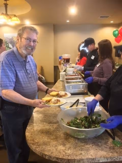 An elderly man in a purple striped polo shirt is smiling and holding a plate of food at a buffet table. Several people wearing gloves are serving food from trays and a large bowl of salad. The setting appears to be a dining area with warm lighting and festive balloons in the background.