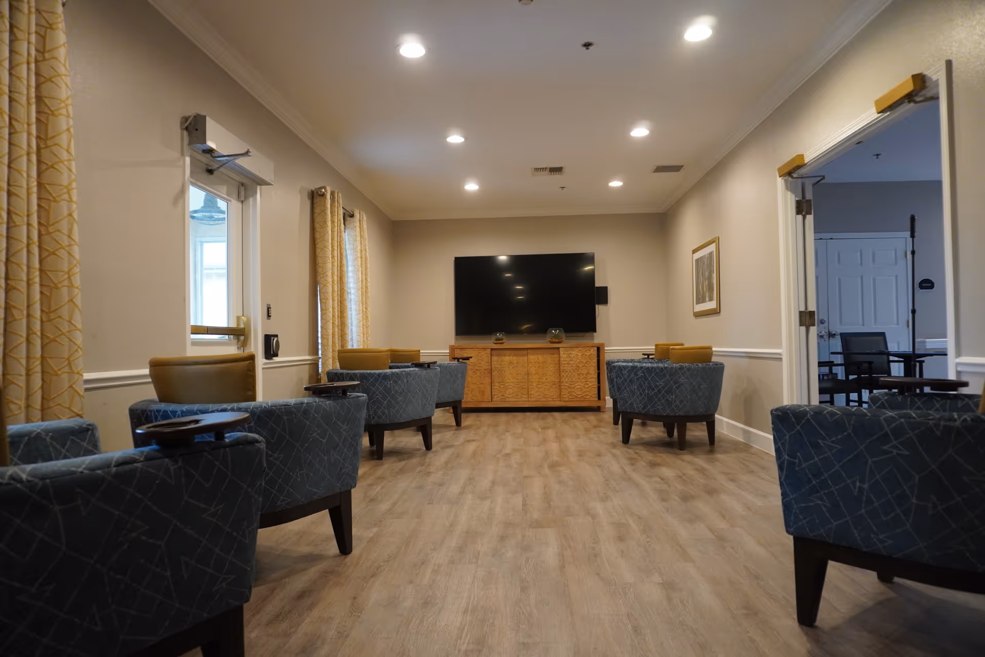A well-lit common seating room with upholstered chairs arranged facing a wall-mounted TV and wooden console.