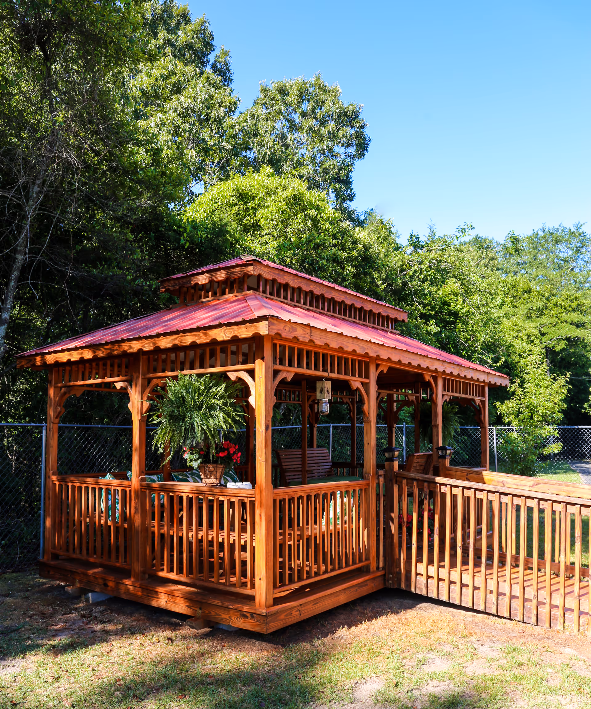 A wooden gazebo with a red roof situated outdoors on grass, surrounded by green trees and a chain-link fence. The gazebo has a ramp for accessibility and contains hanging plants and wooden benches inside.