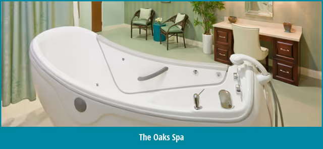 A modern spa room featuring a large white spa tub with built-in controls and a handrail. In the background, there are two chairs with cushions, a small table with a plant, and a vanity desk with a chair and decorative items. The room has soft green walls and curtains.