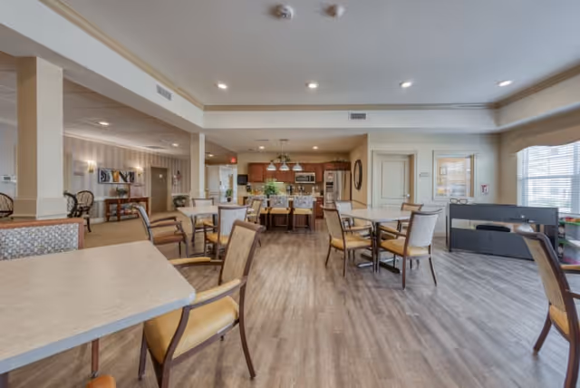 A spacious and well-lit dining area in a senior living facility with multiple tables and chairs arranged on a wood floor. In the background, there is a kitchen area with wooden cabinets, a refrigerator, and a microwave. Large windows on the right side allow natural light to fill the room.