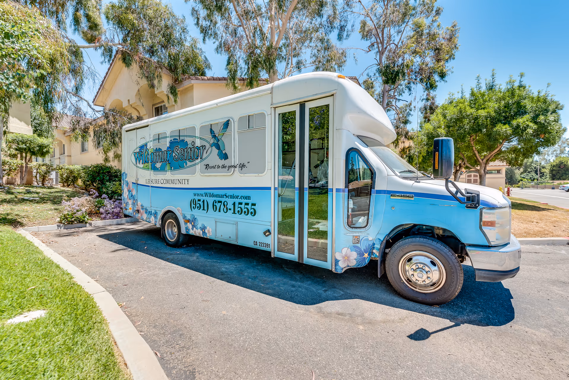 Light-blue Wildomar Senior shuttle bus parked outside a senior living building under trees.