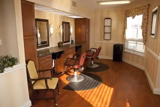 Interior view of a salon area in a senior living facility with two salon chairs in front of a counter with mirrors and lighting. There is a window with floral curtains letting in natural light, a small chair to the side, and wooden flooring throughout.