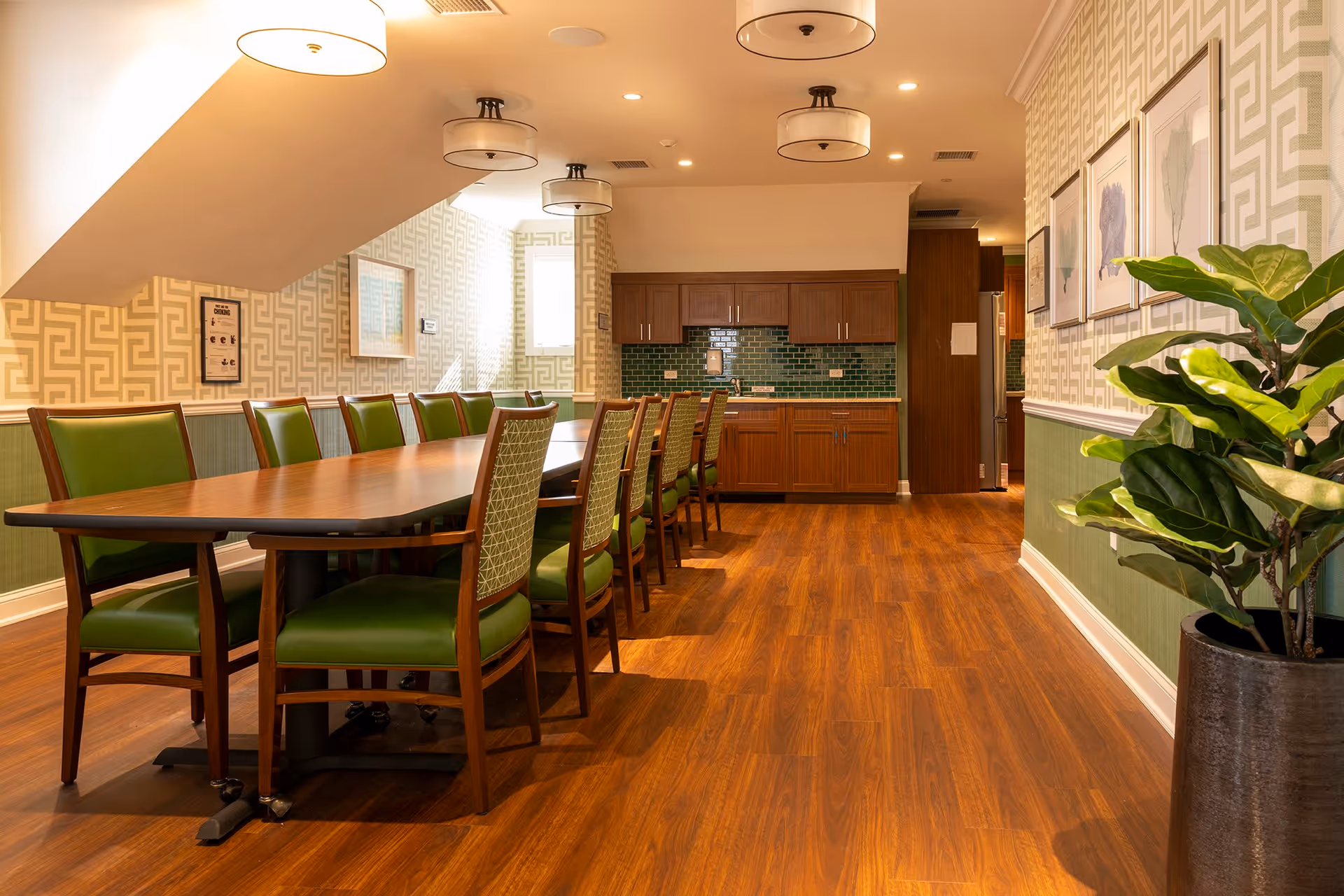 A long wooden dining table with green cushioned chairs arranged around it in a well-lit room with patterned wallpaper and wooden flooring. The room features a kitchenette with wooden cabinets and green tiled backsplash at the far end. There are framed artworks on the wall and a large potted plant in the foreground on the right.