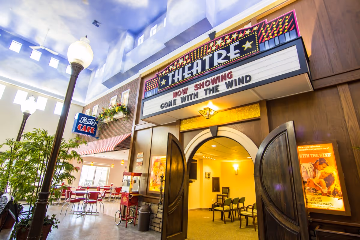 Interior view of a senior living facility featuring a vintage-style theatre entrance with a marquee reading 'THEATRE NOW SHOWING GONE WITH THE WIND'. The theatre doors are open, revealing chairs inside. To the left, there is a cafe area with red chairs and tables, a Pepsi-Cola cafe sign, and decorative plants under a ceiling painted to resemble a blue sky with clouds and windows.