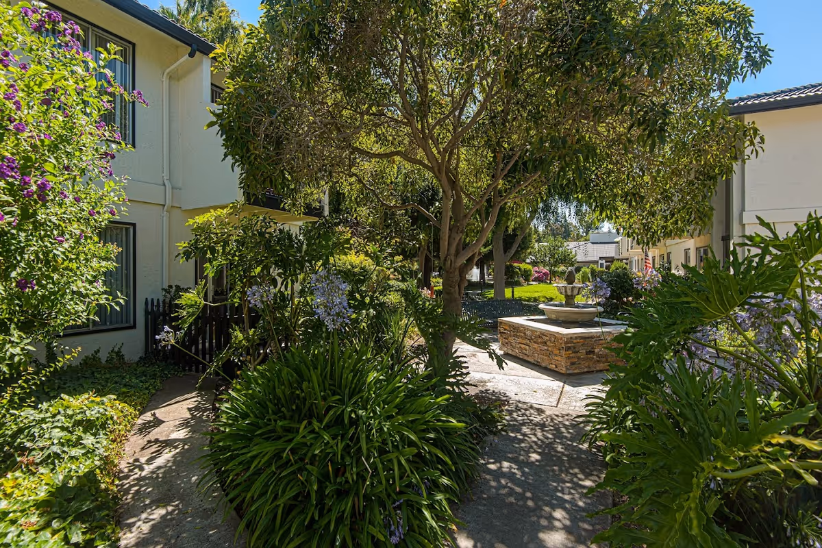 A sunny outdoor garden area at Villa Serena Retirement Community featuring a stone water fountain, lush green plants, trees, and flowering bushes. The garden is surrounded by light-colored buildings with windows and a pathway leading through the greenery.