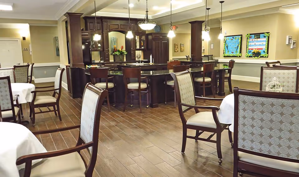Dining room with round tables, upholstered chairs, and a central wooden bar area under pendant lights.