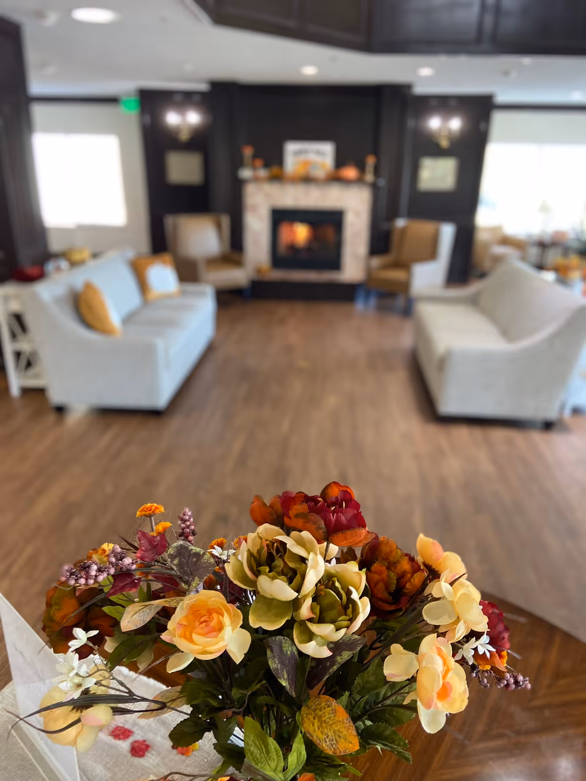 A cozy senior living room with a bouquet of colorful artificial flowers in the foreground. In the background, there are two light-colored sofas with yellow pillows, two armchairs, and a fireplace with a lit fire. The room has wooden flooring and dark wood paneling around the fireplace.