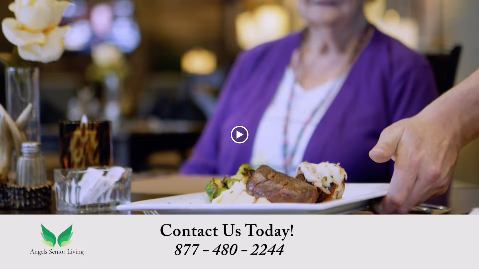 A close-up of a hand serving a plate with a steak, mashed potatoes, and vegetables to an elderly woman wearing a purple cardigan seated at a dining table in a senior living facility.