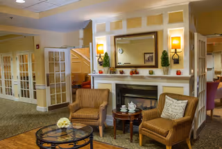 A cozy seating area in a senior living facility featuring two cushioned armchairs with a small round table between them holding a teapot and cups. Behind the chairs is a white fireplace with a large mirror above it, flanked by two wall sconces with warm lighting and small decorative plants. To the left, there are glass-paneled double doors and a staircase in the background.