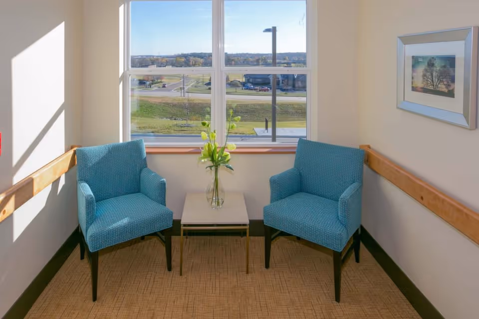 A small seating area with two blue upholstered armchairs facing each other with a small square table in between. On the table is a clear vase with green and yellow flowers. The seating area is next to a large window showing an outdoor view of a road, grassy area, and distant trees. There is a framed picture on the wall to the right and wooden handrails along the walls.