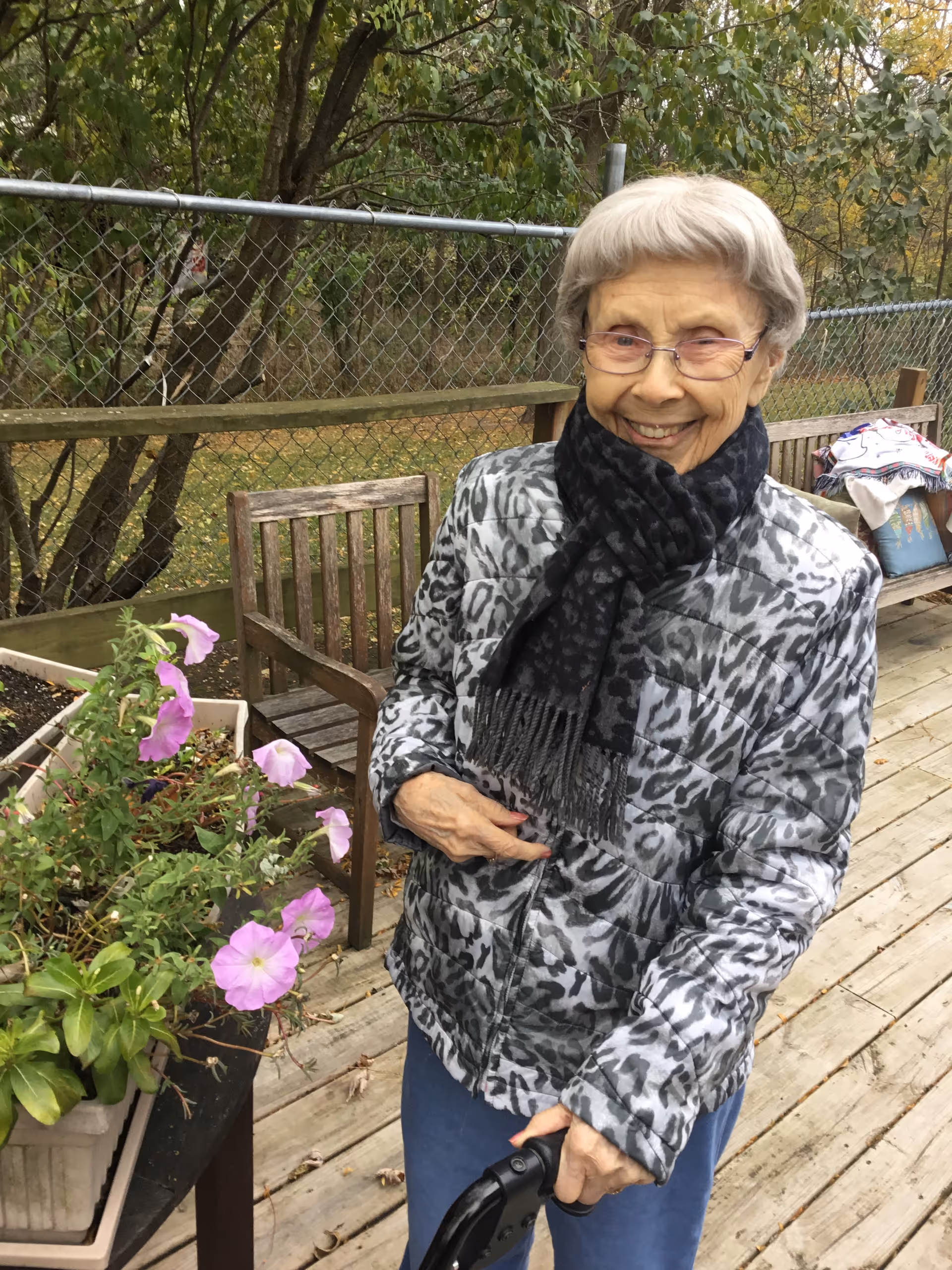 An elderly woman wearing glasses, a gray patterned jacket, and a black scarf is standing on a wooden deck outdoors. She is smiling and holding a black cane. Behind her, there is a wooden chair, a planter with pink flowers, and a chain-link fence with trees and greenery in the background.