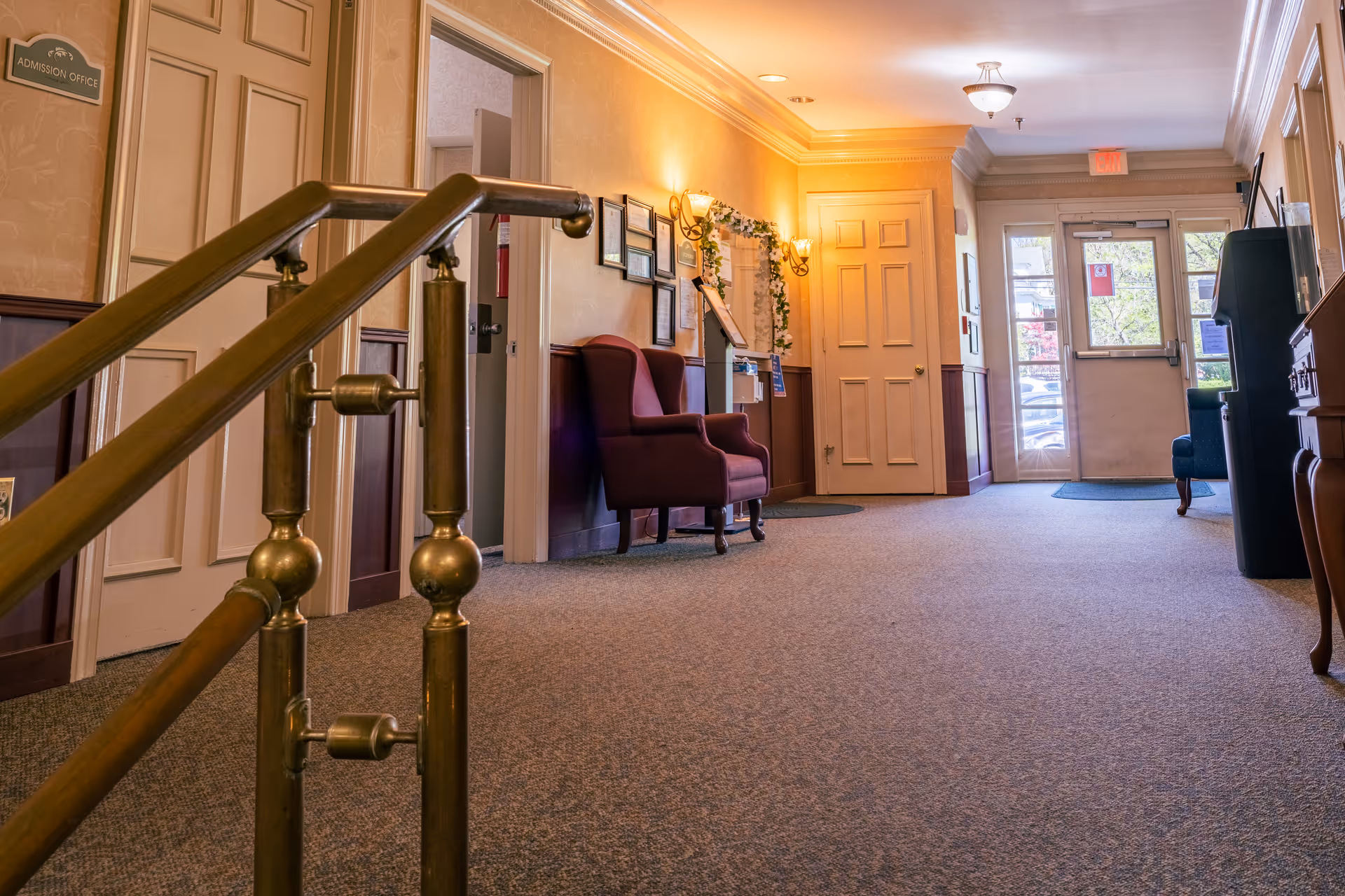 Interior hallway of a senior living facility with carpeted floor, brass handrails, wall sconces, framed pictures, and upholstered chairs. A door labeled 'Admission Office' is visible on the left, and a glass exit door is at the far end of the hallway.
