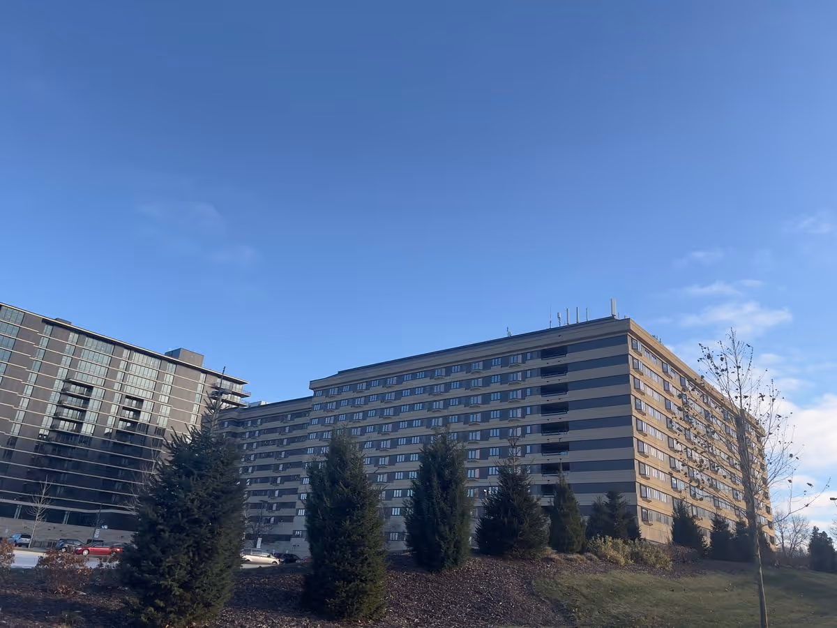 Low-angle view of a multi-story apartment building with a striped facade, landscaped evergreens in front, and a clear blue sky.